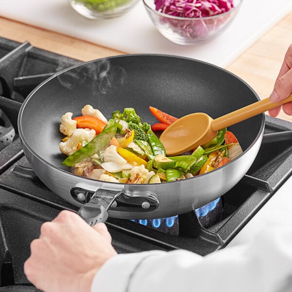 A person stirring food in an Emperor's Select aluminum stir fry pan on a gas stove.