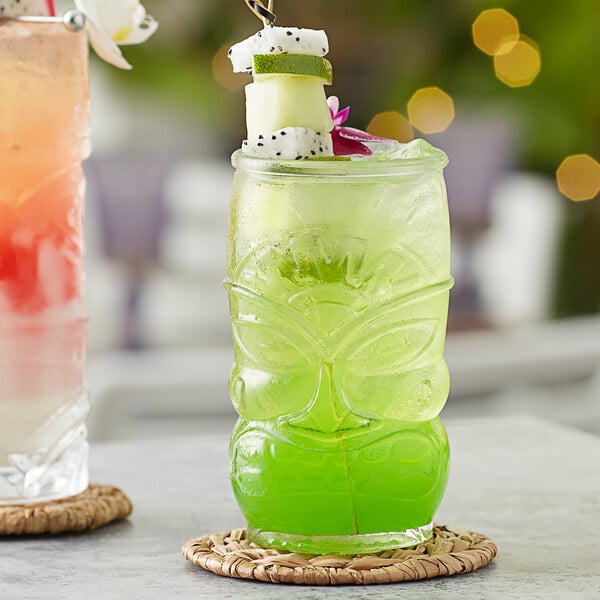 A green Acopa Tiki glass filled with a green tropical drink with a tropical fruit garnish on top, on a table.