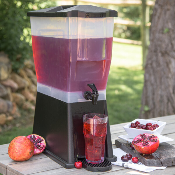 A Carlisle black plastic beverage dispenser with red liquid, ice, and a cherry, with a bowl of cherries and a pomegranate on the counter.