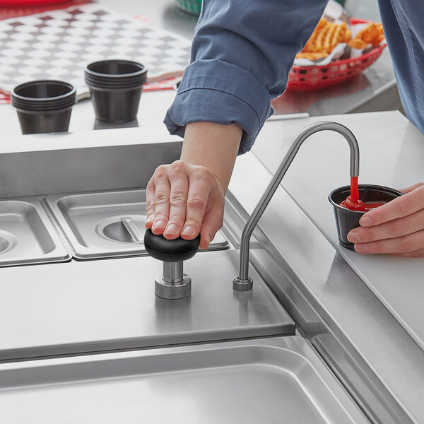 A hand using a ServSense stainless steel pump to pour ketchup into a black plastic cup on a white surface.