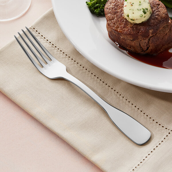 An Acopa Triumph stainless steel dinner fork next to a plate of food with a piece of meat and butter.
