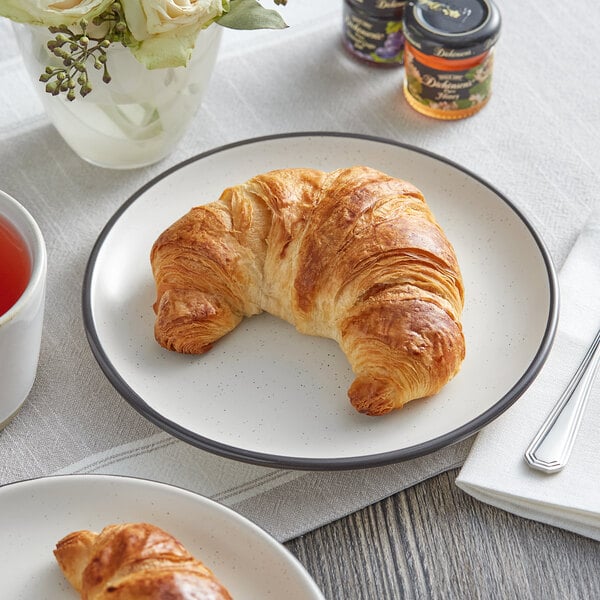 A Schulstad large curved butter croissant on a plate next to a cup of tea.