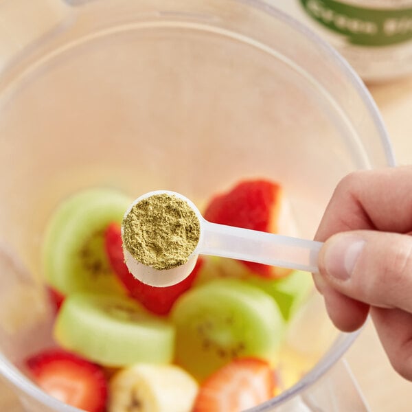 A hand holds a measuring spoon full of Add A Scoop Green Blend supplement powder over a bowl of fruit.