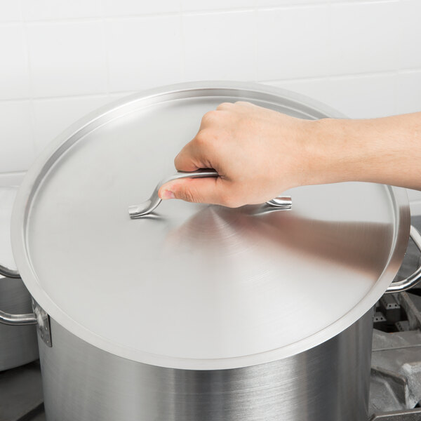 A hand holding a Vollrath metal lid over a silver pot.