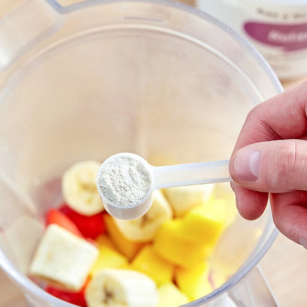 A hand holding a measuring spoon of Add A Scoop Relaxing Blend supplement powder over a blender with fruit.
