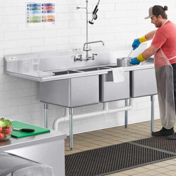 A man wearing gloves and an apron washing dishes in a Regency stainless steel three compartment sink.