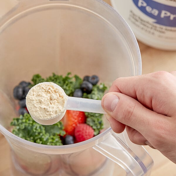 A hand holding a scoop of Add A Scoop Pea Protein powder over a blender.