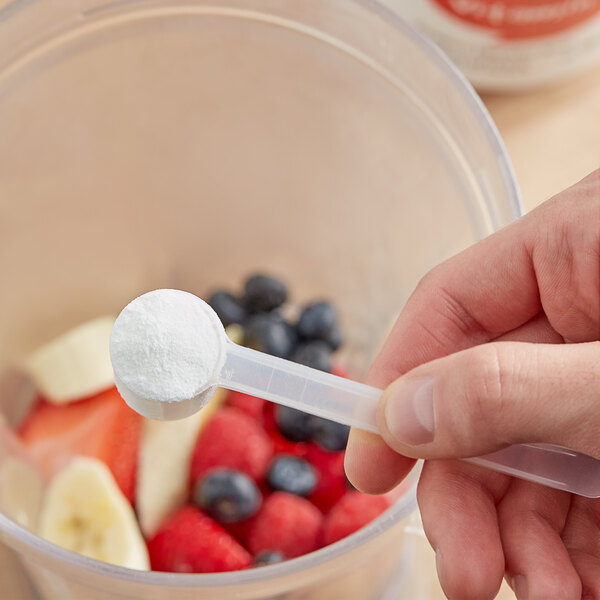 A hand using a spoon to add Add A Scoop Vitamin C Blend supplement powder to a bowl of fruit.