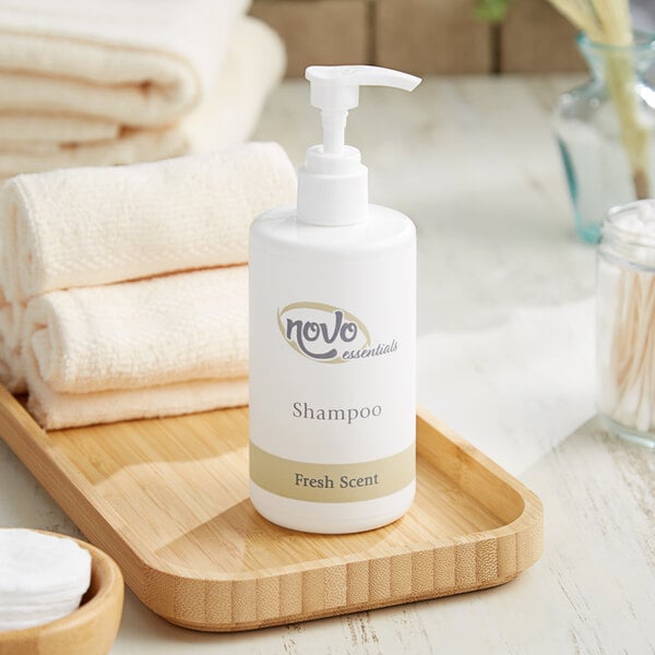 A white bottle of Novo Essentials Hotel and Motel Shampoo on a wooden tray next to white towels.