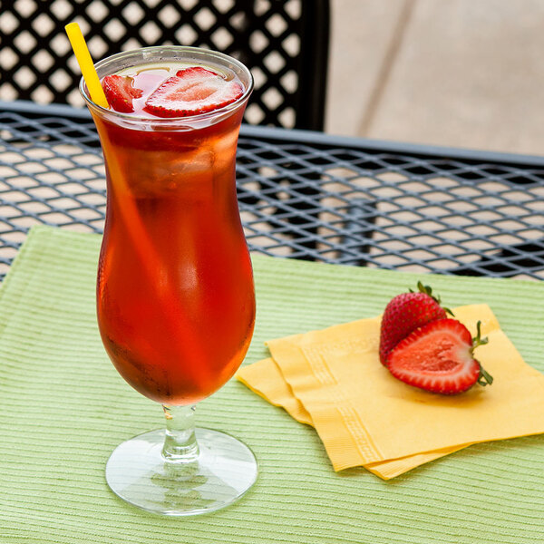 A Libbey hurricane glass of red liquid with strawberries and a straw on a napkin.