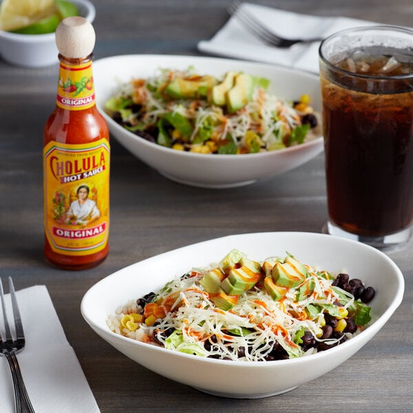 A bowl of salad on a table with a bottle of Cholula Original Hot Sauce.