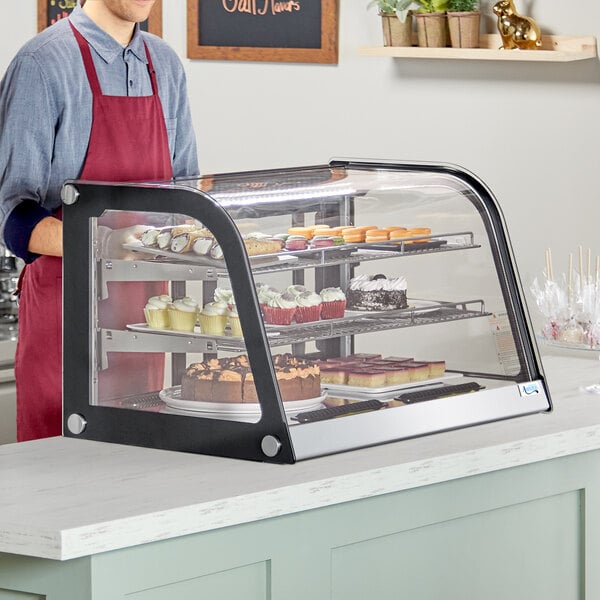 An Avantco curved refrigerated countertop bakery display case on a counter with food in it and a man wearing a red apron standing behind it.