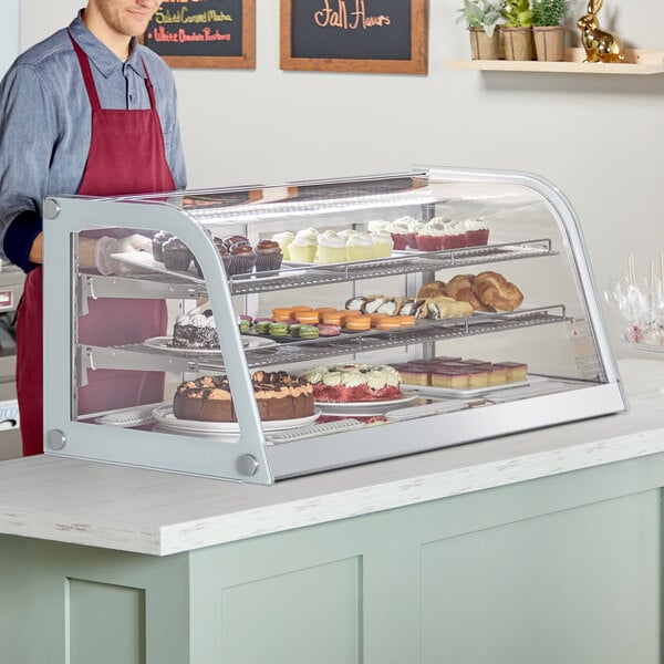 A man standing behind a white Avantco countertop bakery display case filled with pastries and cupcakes.