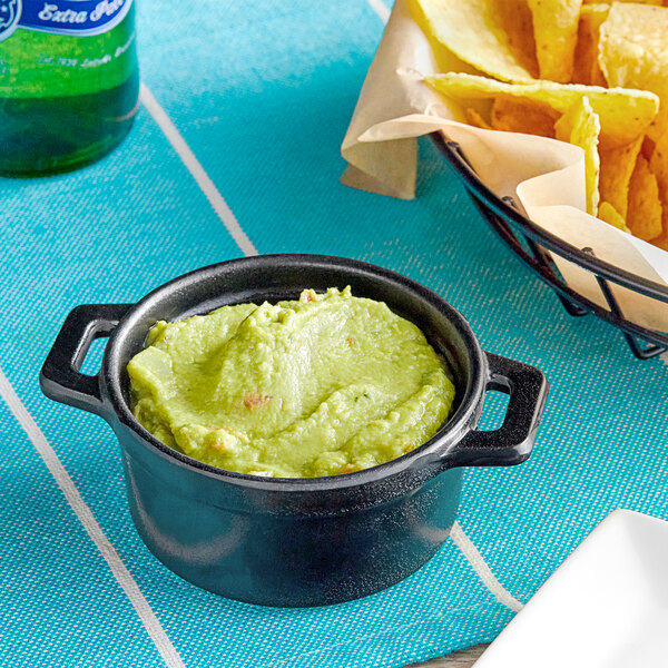 An Elite Global Solutions black faux cast iron melamine pot filled with guacamole on a table with blue tablecloth and a bowl of chips.