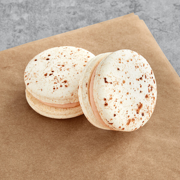 Two Macaron Centrale Amaretto macarons with white chocolate coating on a brown surface.
