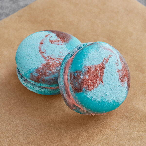 Two blue and pink Macaron Centrale macarons with blue and red icing on a brown surface.