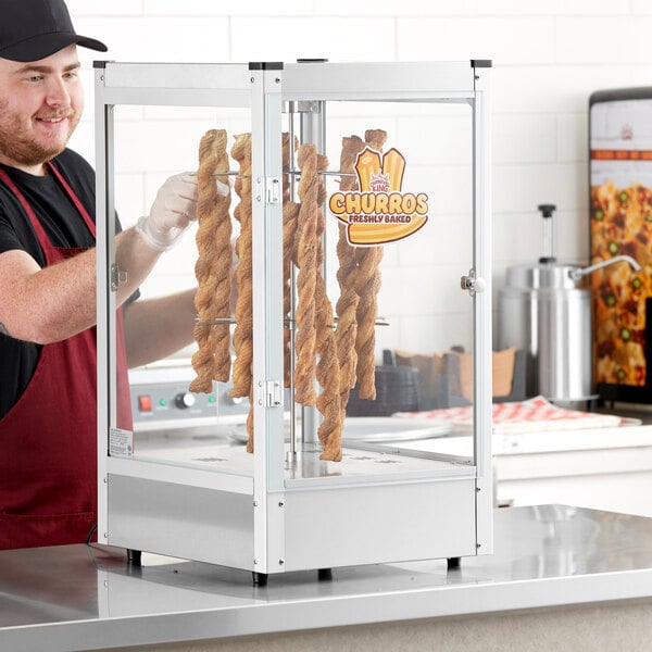 A man in gloves and a black shirt uses a Carnival King churro display warmer to hold churros in a glass case.