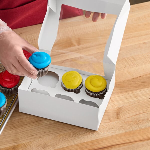 A person wearing gloves puts a yellow frosted cupcake into a white Baker's Mark cupcake box.