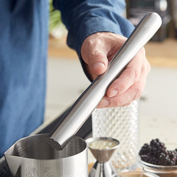 A person holding an Acopa stainless steel muddler over a metal container with a stick.