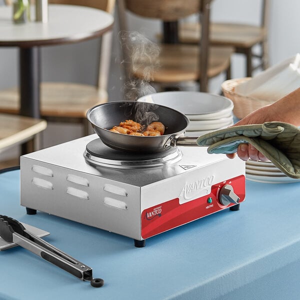 A hand holding a pan of shrimp cooking on an Avantco portable electric hot plate.