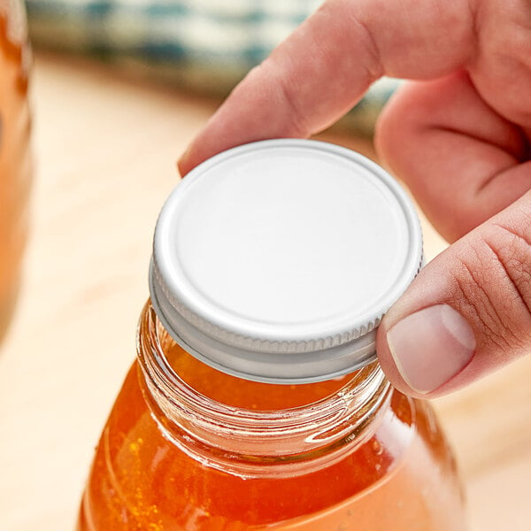 A person's hand holding a jar with a white metal lid containing orange liquid.