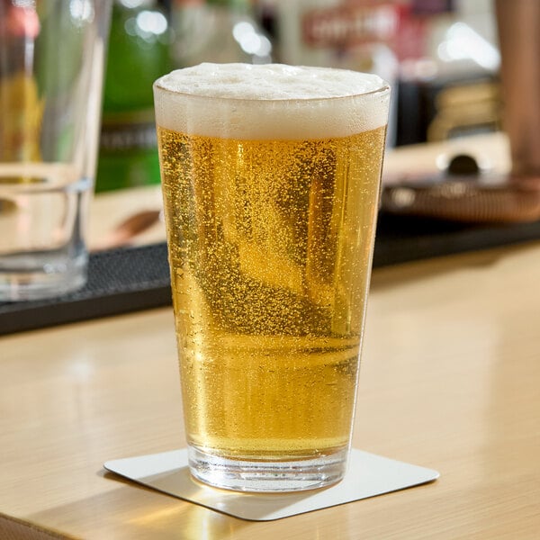 A clear 16 oz. stackable cooler or mixing glass filled with a light, bubbly beverage and topped with foam, placed on a coaster.