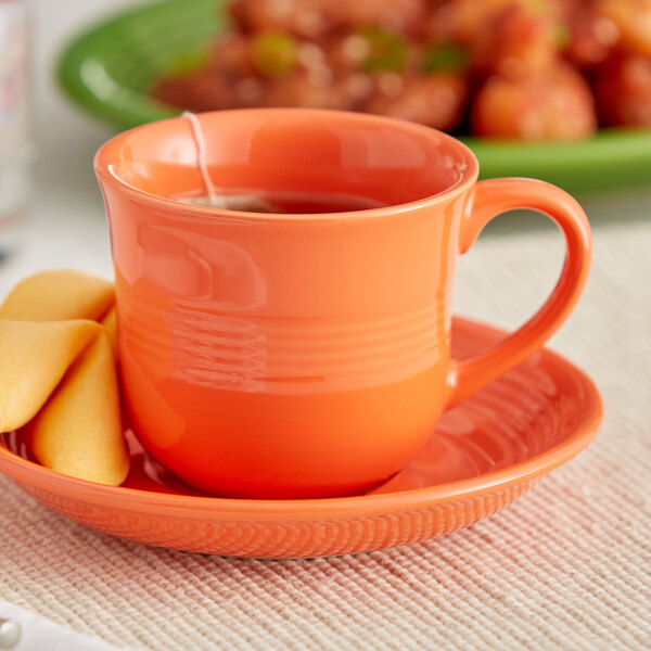 An Acopa Valencia orange stoneware cup filled with tea on a saucer with a slice of lemon.