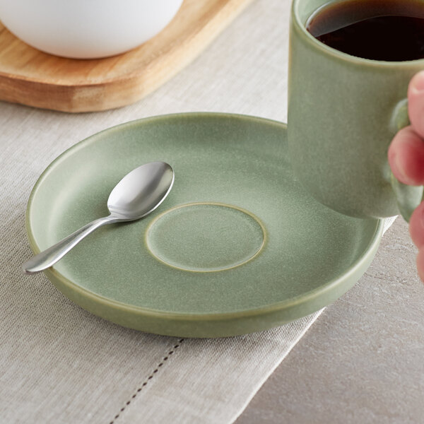 A hand holding a cup of coffee and a spoon on a sage matte porcelain saucer.