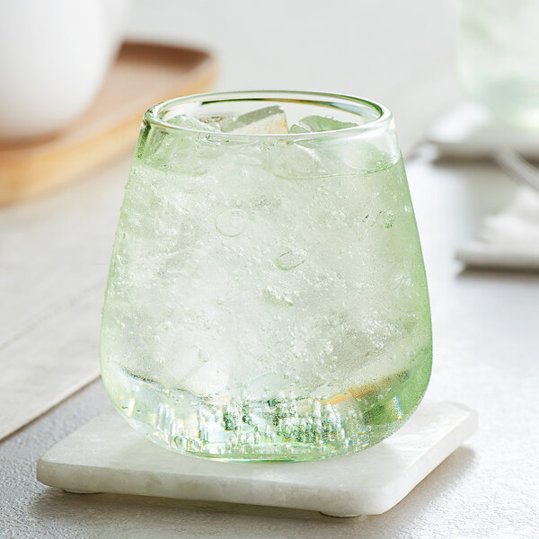A green Acopa Pangea stemless wine glass filled with clear liquid and ice on a white marble coaster.