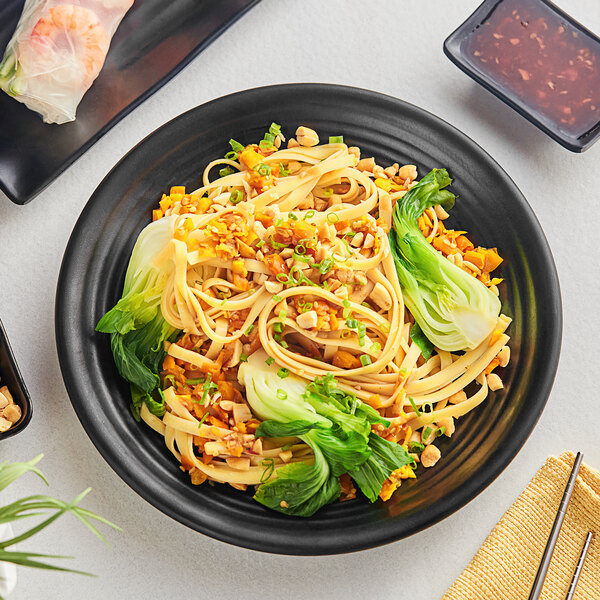A black Acopa Izumi coupe plate with noodles and vegetables and a pair of chopsticks.