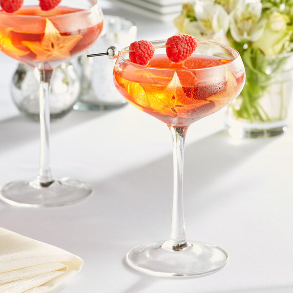 A pair of Acopa Select coupe glasses with orange liquid and raspberries.