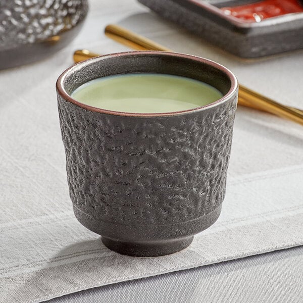 An Acopa Heika black stoneware tea cup filled with green liquid on a table.