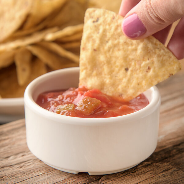 A person holding a chip and dipping it into a Carlisle ivory ramekin of salsa.