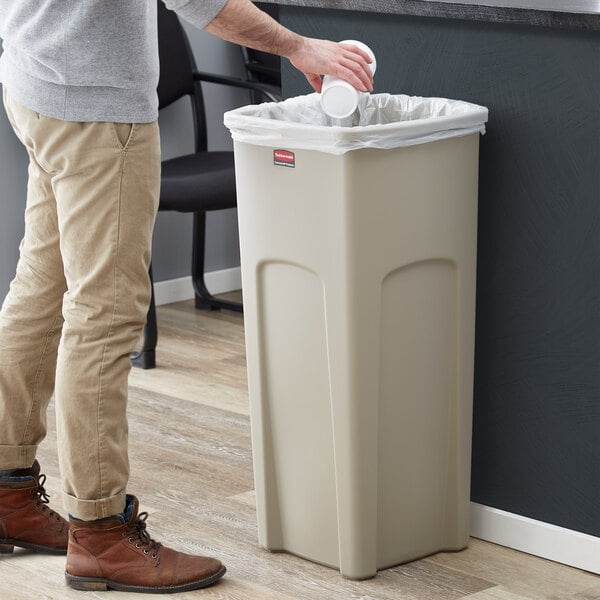 A person standing next to a Rubbermaid beige square trash can.
