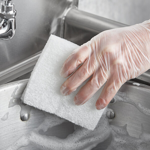 A hand in a plastic glove using a white Lavex multi-purpose sponge to clean a sink.