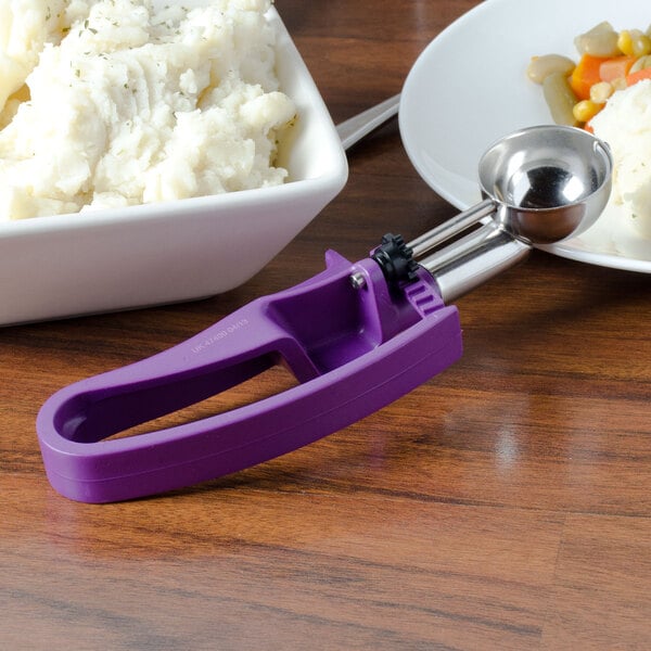A white bowl of mashed potatoes with a Vollrath purple handled disher on a plate.