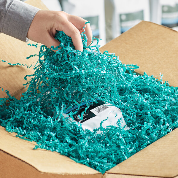 A hand holding a package of teal Spring-Fill crinkle cut paper in a cardboard box.