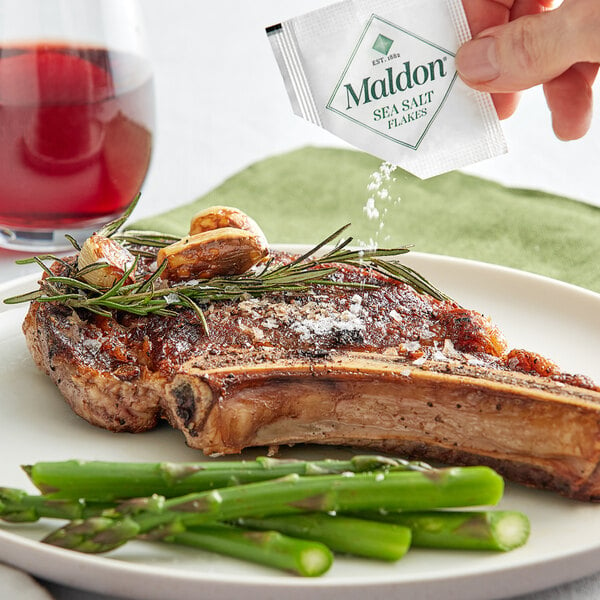 A hand sprinkling Maldon Sea Salt on a piece of meat with rosemary and garlic.