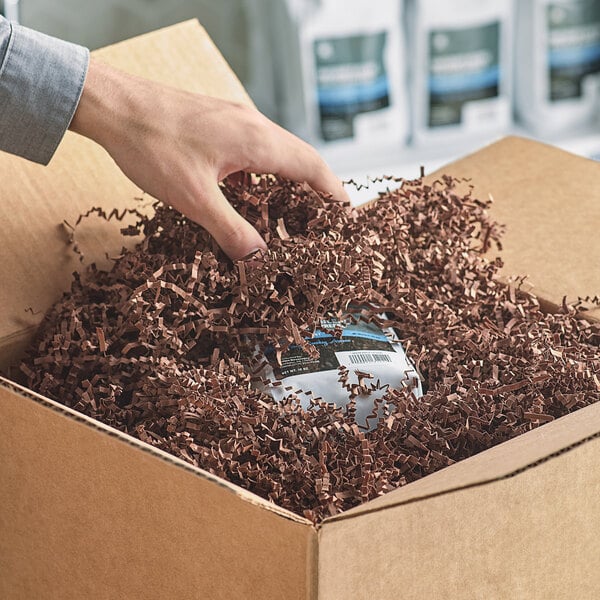 A hand reaching into a cardboard box filled with brown Spring-Fill Chocolate Crinkle Cut paper shred.