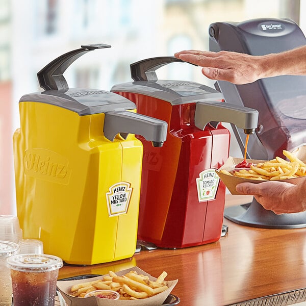 A person using a red and yellow Heinz pump to add ketchup to a bowl of french fries.