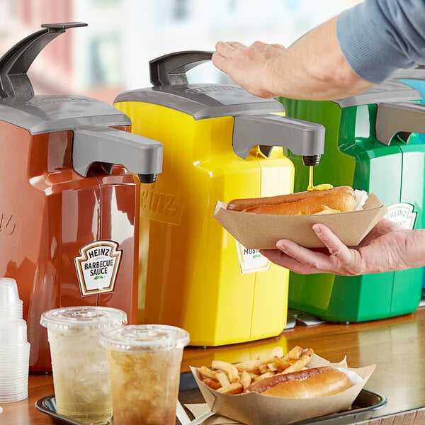 A person using a Heinz countertop pump to add yellow mustard to a hot dog in a plastic cup.