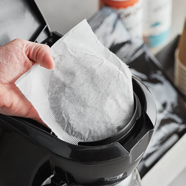A hand holding a round white paper filter over a black coffee maker.