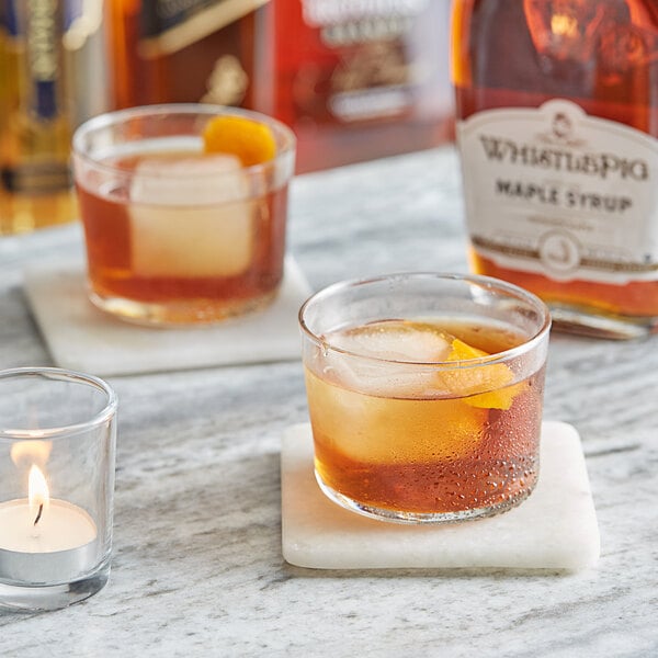 A bottle of Runamok WhistlePig Rye Whiskey Barrel-Aged Maple Syrup on a table with two glasses of brown liquid and ice with a candle.