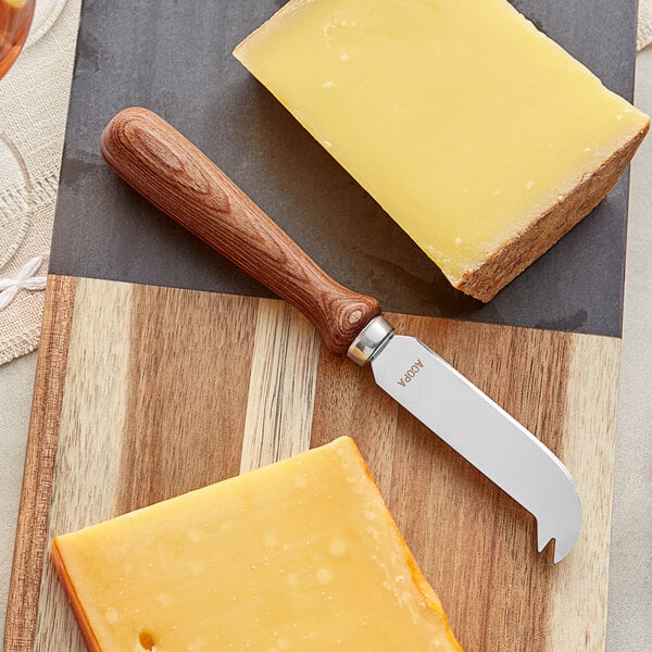 An Acopa stainless steel cheese knife next to a block of cheese on a cutting board.