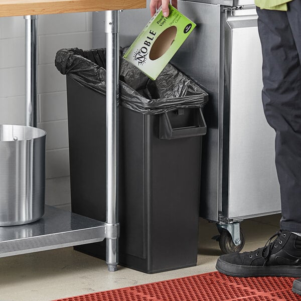 A person standing next to a Lavex slim black rectangular under-counter trash can with a green bag in a kitchen.