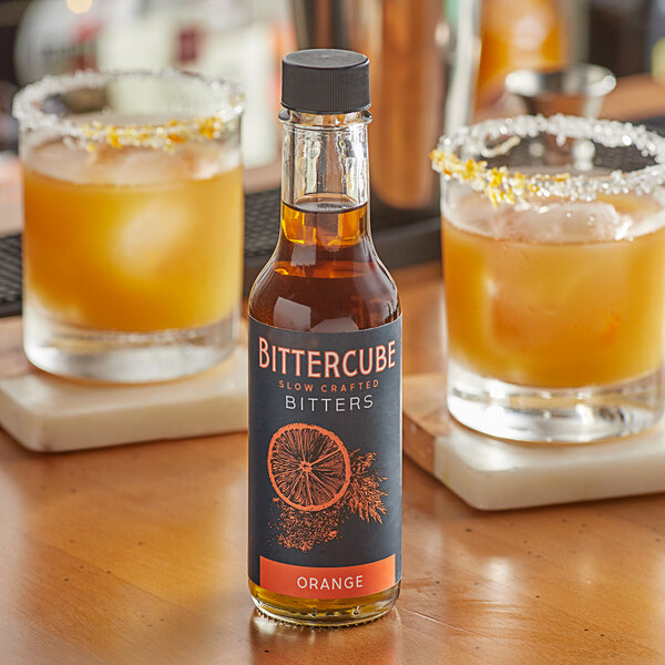 A bottle of Bittercube Orange Bitters on a table with glasses of liquid.