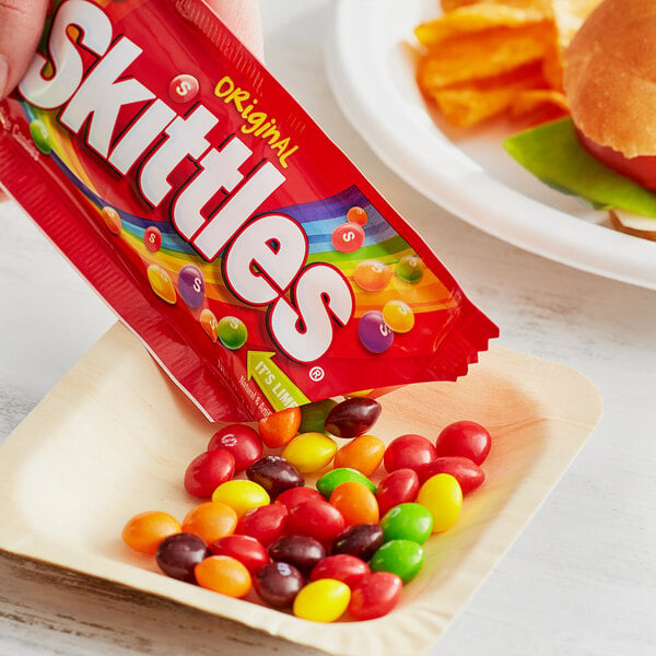A hand holding a SKITTLES® Original Fruity Candies pouch over a plate of candy.
