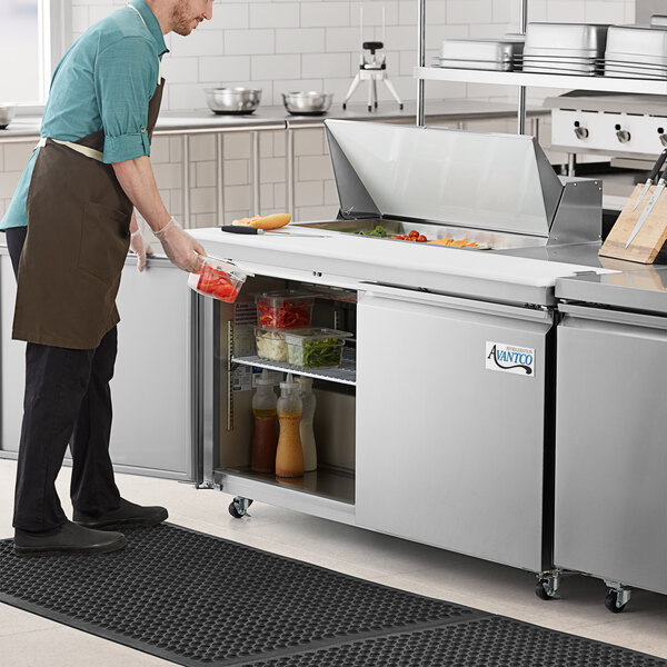 An Avantco stainless steel sandwich prep table in a commercial kitchen with a man wearing a brown apron and gloves.