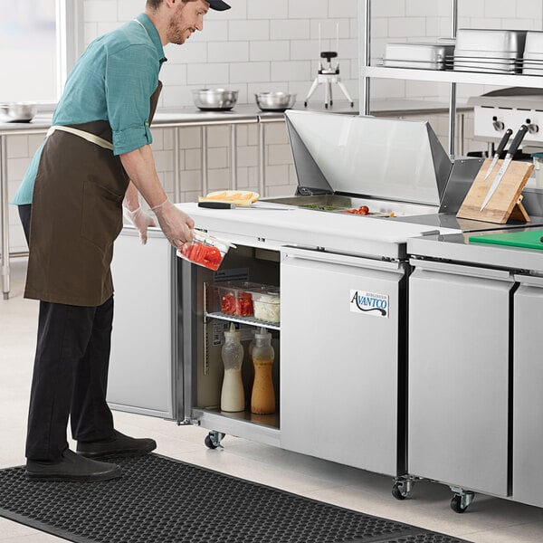 An Avantco stainless steel sandwich prep table on a white surface with a container of food.