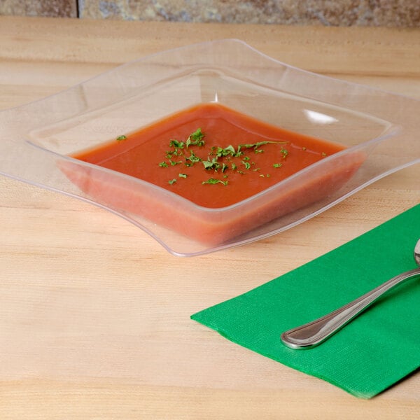 A Fineline clear plastic bowl of tomato soup with a spoon on a table.